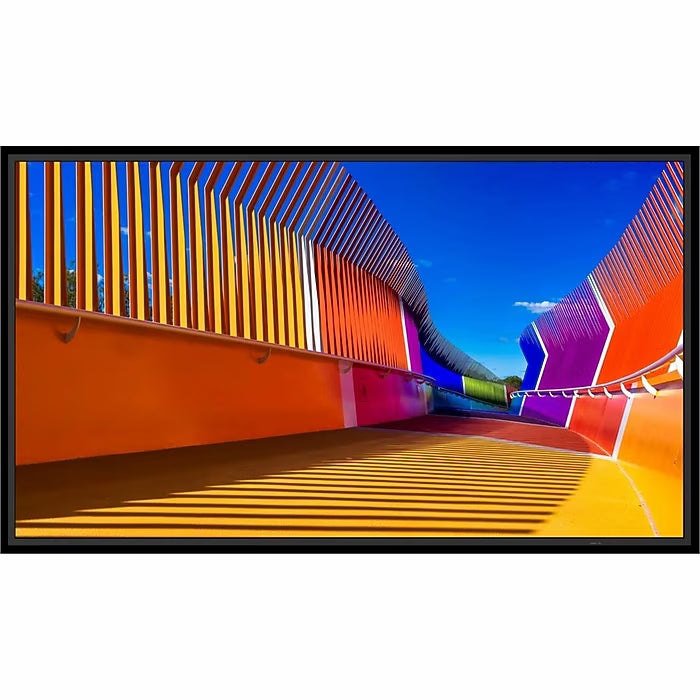 A Sharp 50″ 4K UHD Professional Display showing a vibrant, multicolored pedestrian bridge with curved walls and slatted railings against a blue sky.